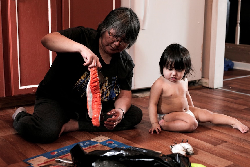 A woman is pictured sitting on the ground while holding up a skinned fish, while a young child sits next to her.