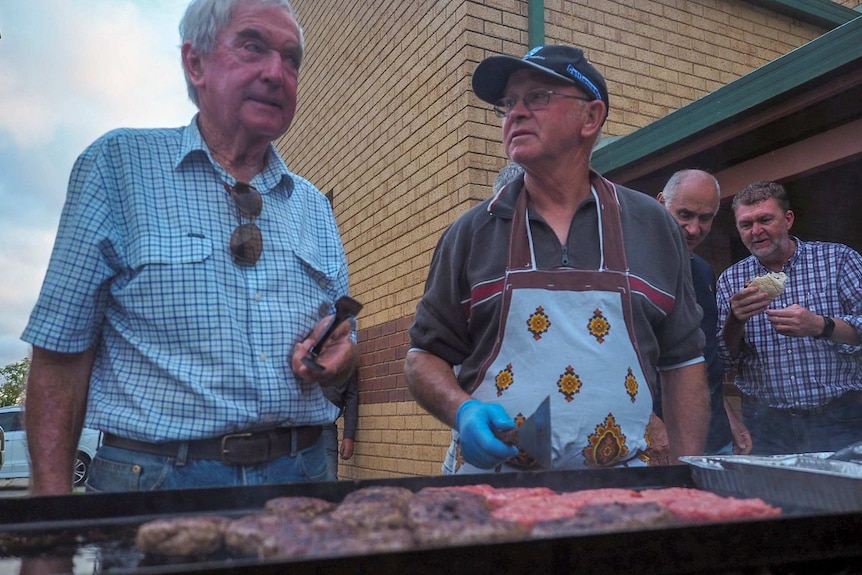 Two men stand talking at a barbecue with one holding tongs and another wearing an apron and holding an egg flip.