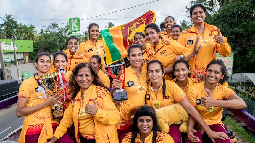 Sixteen happy women holding Sri Lankan flags and trophies on a roofless bus driving through streets.