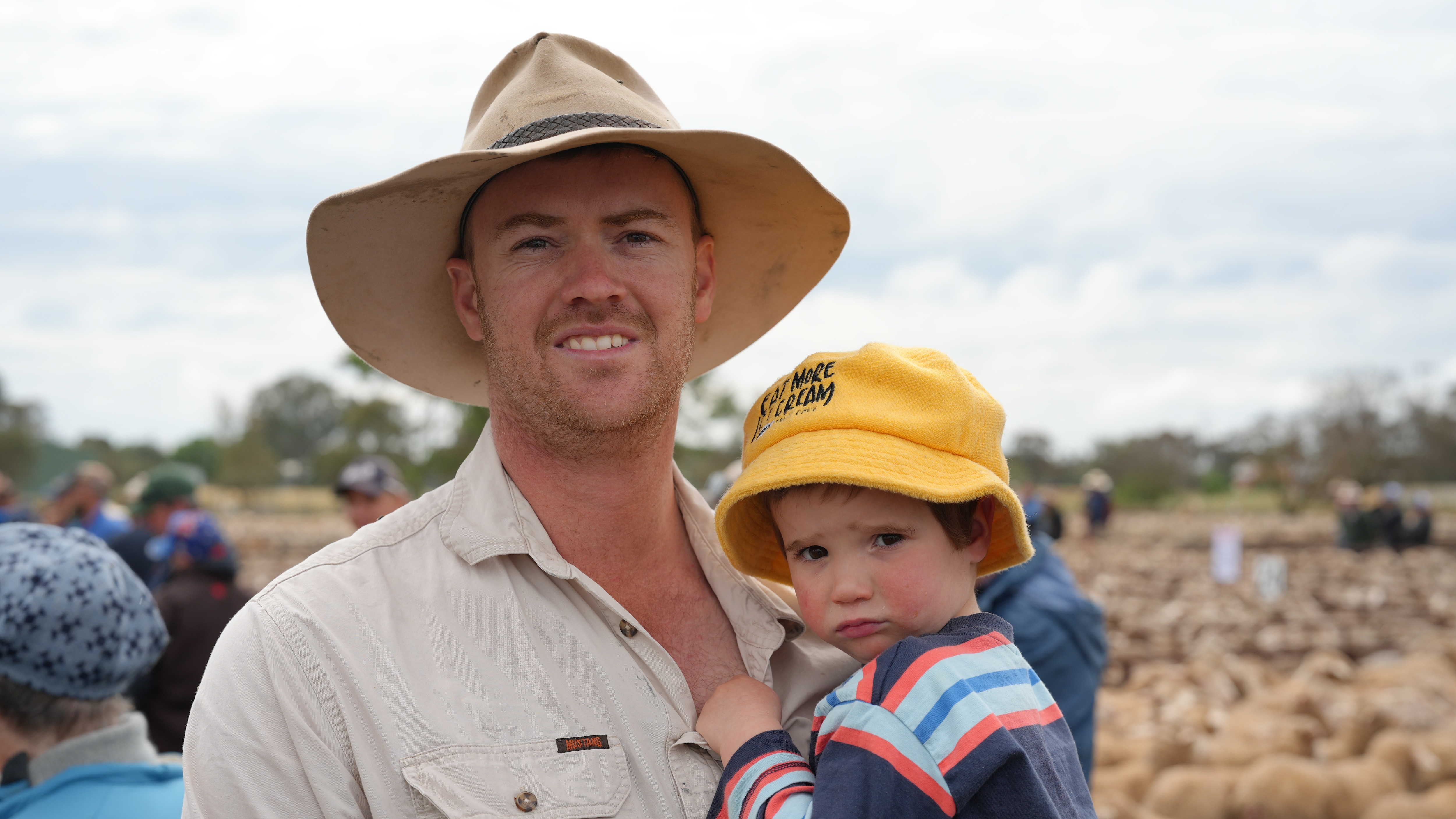 A man in a hat holding a toddler with sheep in the background.