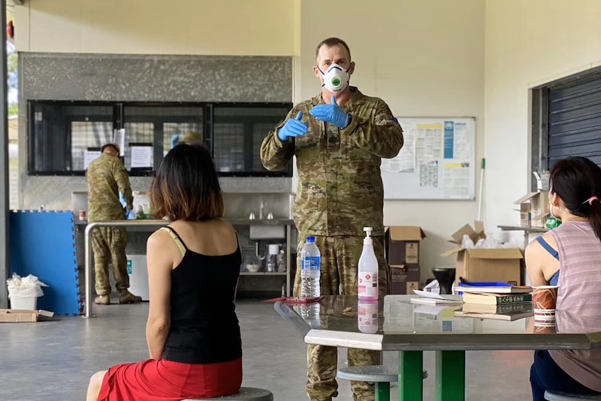 A soldier wearing a face mask and blue gloves stands in front of a table talking to evacuees.