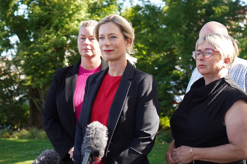 Rebecca White flanked by two other women at a press conference, standing before a microphone.