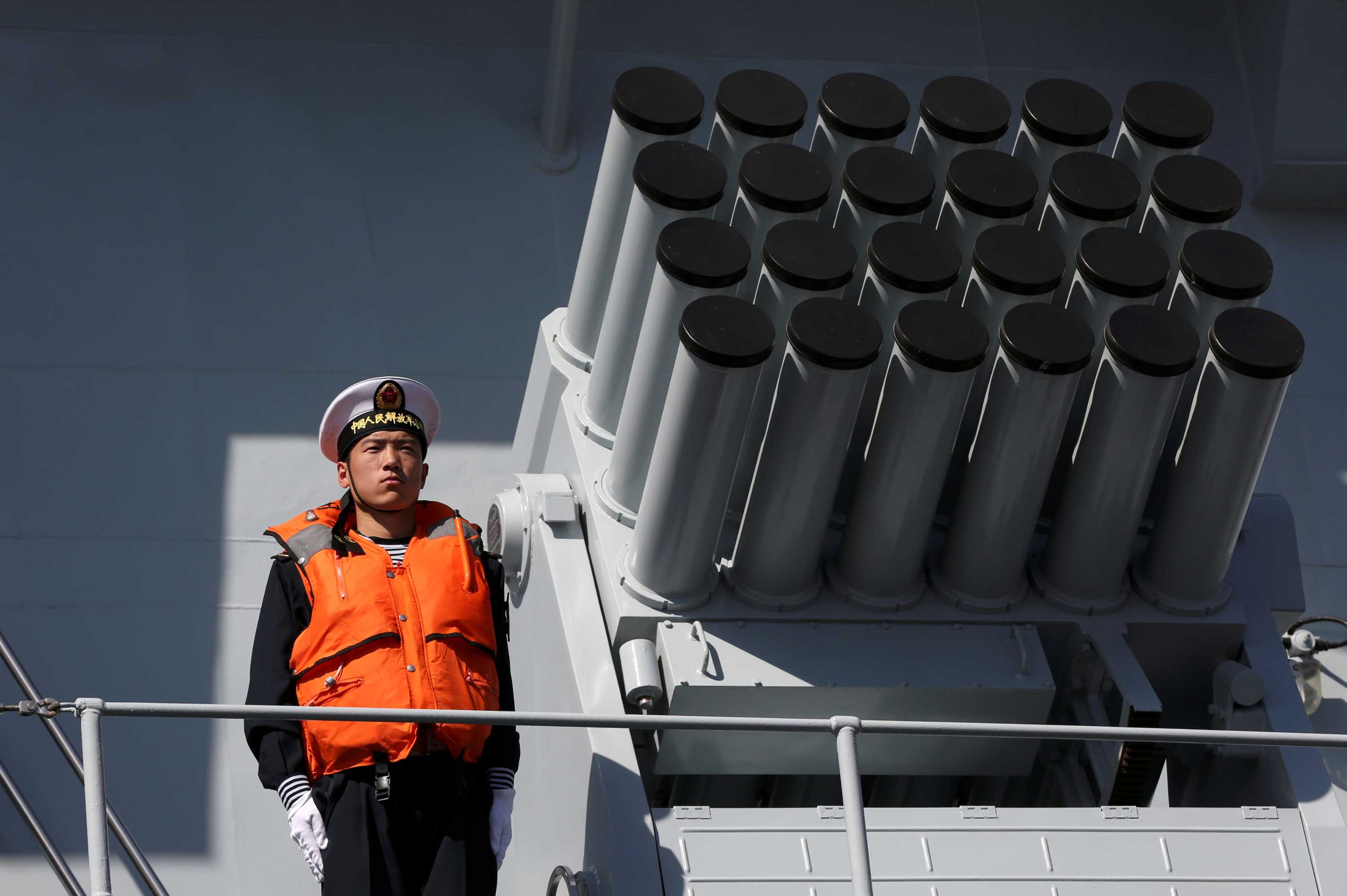 A man in a navy uniform and life jacket stands beside a rocket launcher on a ship.