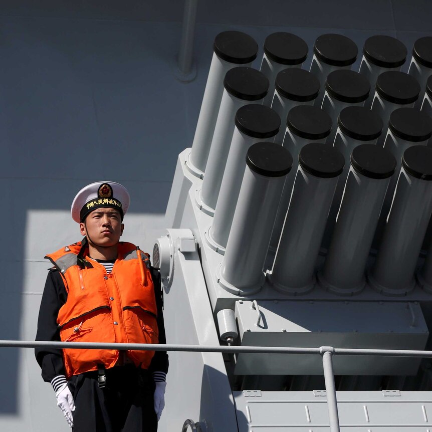 A man in a navy uniform and life jacket stands beside a rocket launcher on a ship.