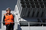 A man in a navy uniform and life jacket stands beside a rocket launcher on a ship.