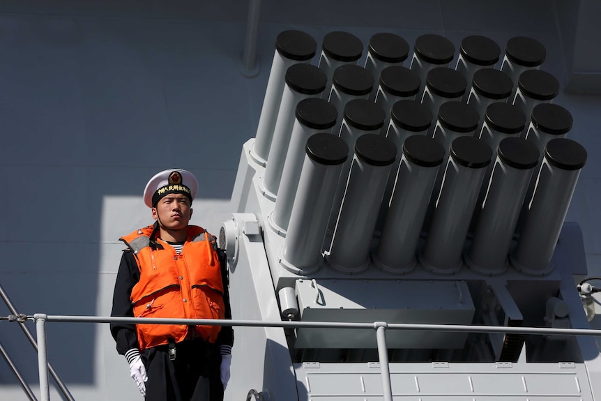 A man in a navy uniform and life jacket stands beside a rocket launcher on a ship.