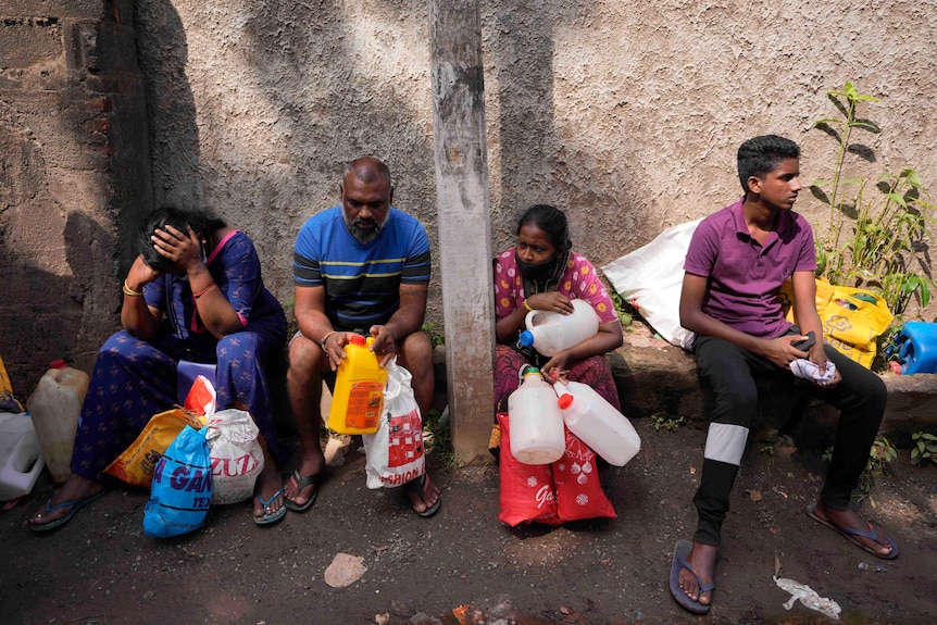 Four people sit on the edge of a kerb holding plastic containers to carry liquid.