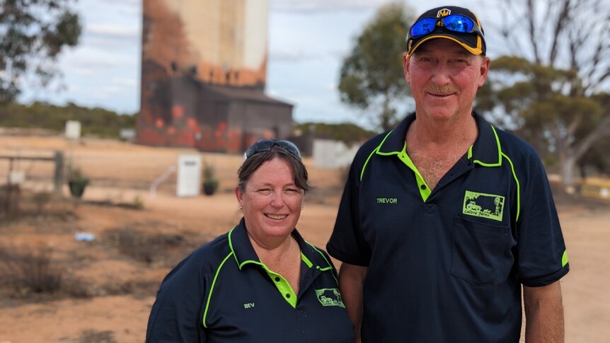 Beverly and Trevor, two white middle-aged people, in navy blue polos stand smiling in front of a painted silo.