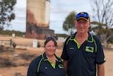 Beverly and Trevor, two white middle-aged people, in navy blue polos stand smiling in front of a painted silo.