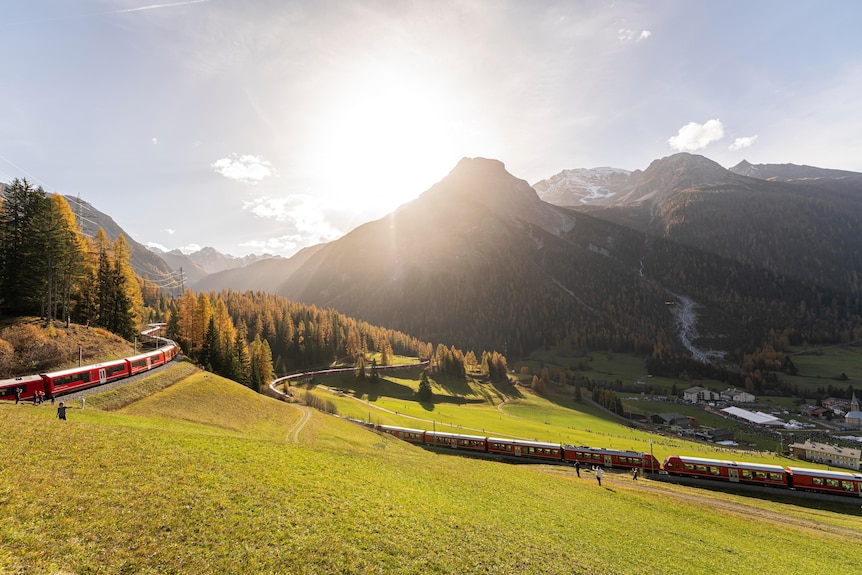 A red train winds around a hill in front of mountains