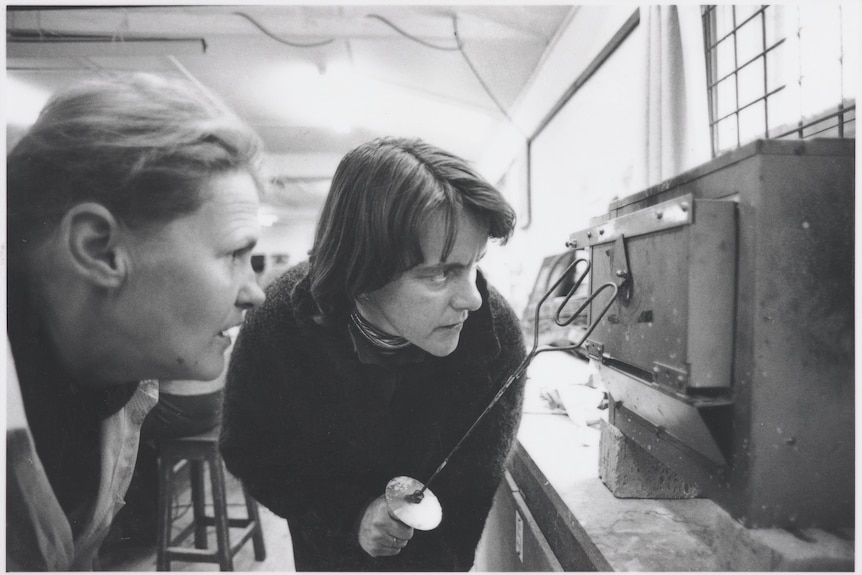 Two women keep a close eye on an oven as part of the enameling process. One of them holds a rounded firing fork.  
