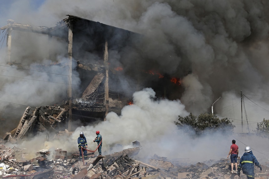 Firefighters and rescue volunteers stand on piles of rubble in front of a smoking, destroyed wreck of a building.