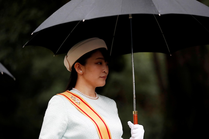 Princess Mako, a young Japanese woman, wears a formal gown, sash, pearl necklace and hat. She is holding a black umbrella