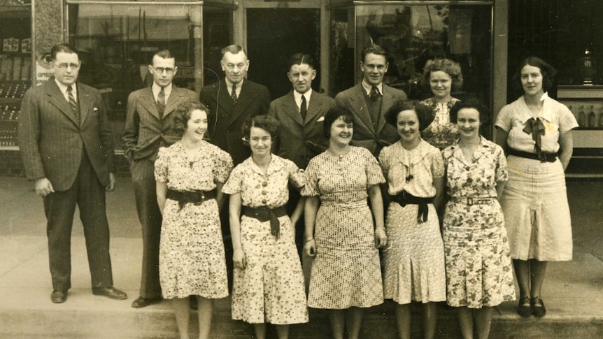 a black and white image of a group of people standing in front of a shop front in 1937