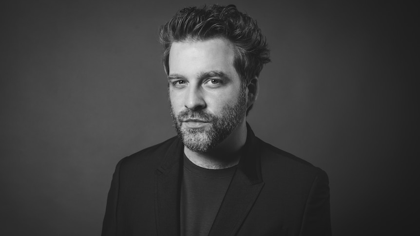 A monochrome photo of pianist Taylor Eigsti in a suit jacket; he's looking at the camera