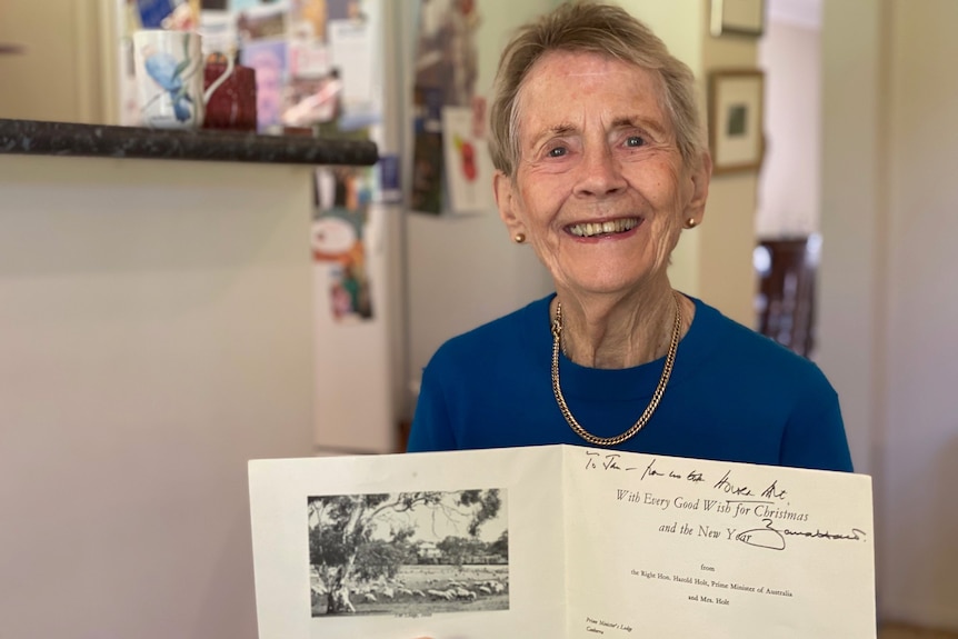 A smiling elderly woman holds open a christmas card with a black and white photo on it.