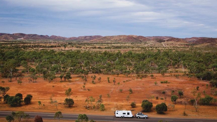 A four-wheel-drive towing a caravan along a bitumen road, captured by drone, surrounded by red dirt and small green shrubs.