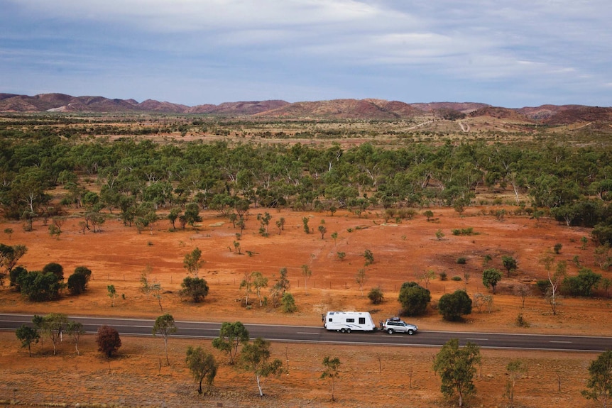 A four-wheel-drive towing a caravan along a bitumen road, captured by drone, surrounded by red dirt and small green shrubs.