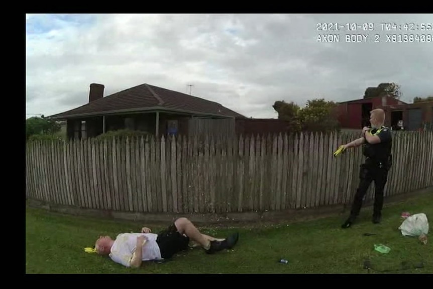 A man lies on some grass in a suburban area while a police officer points a taser at him.
