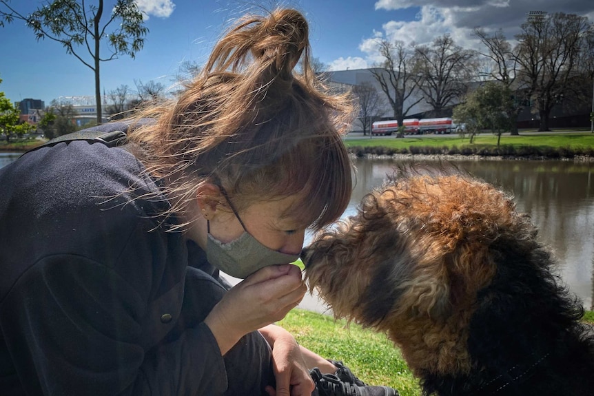 A woman sits along the banks of the Yarra smiling at her brown and black dog.