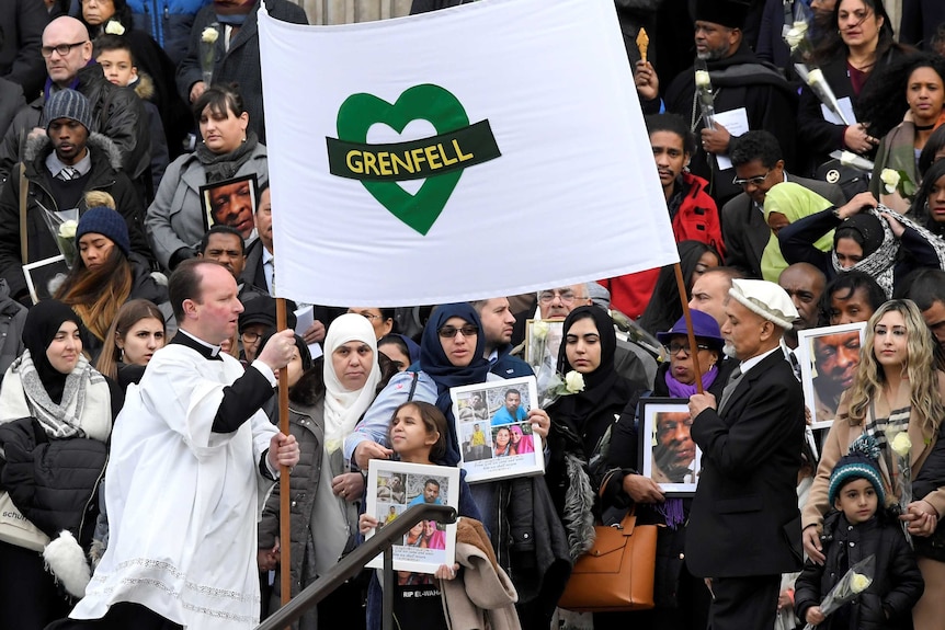 A crowd of people are standing outside holding pictures of their loved ones who died in the fire.