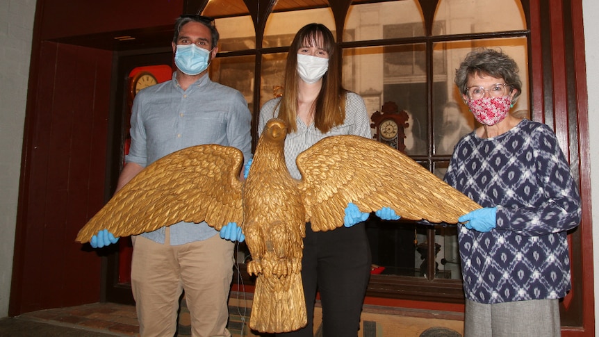 Three people in masks hold large gold sculpture of an eagle in front of a replica of an old clock shop.