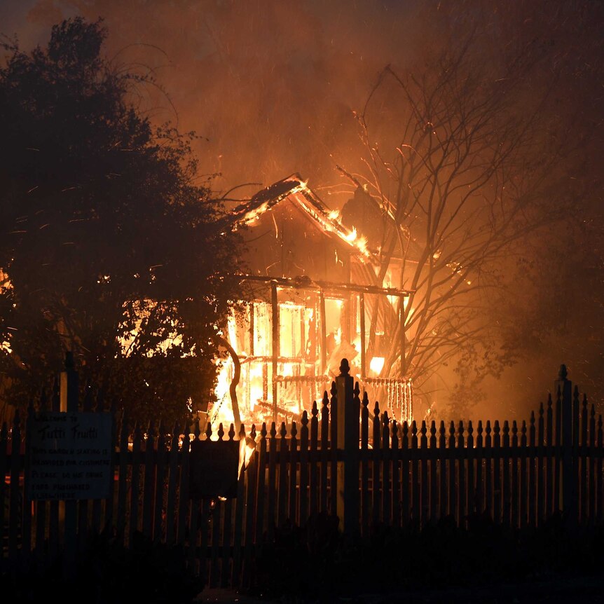 Fire engulfs the front of a house. The front fence is silhouetted by the flames.
