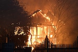 Fire engulfs the front of a house. The front fence is silhouetted by the flames.