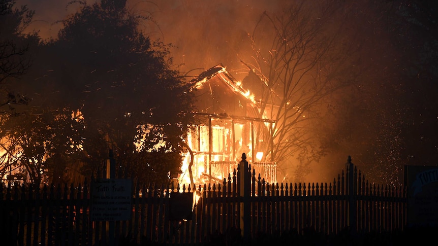 Fire engulfs the front of a house. The front fence is silhouetted by the flames.