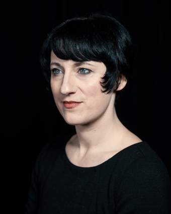 Headshot of woman with louise brooks style dark bob and red lips, against black background.