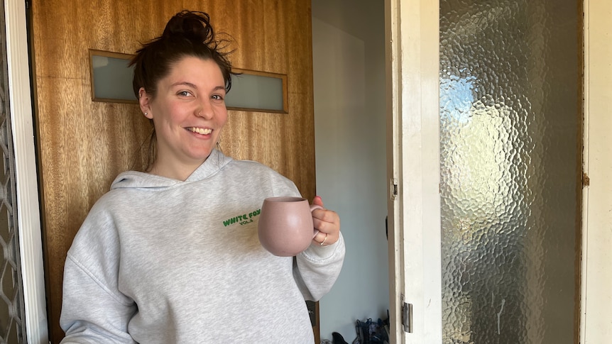 A smiling woman with dark hair in a bun wearing a grey hoodie holding a mug in the front doorway.
