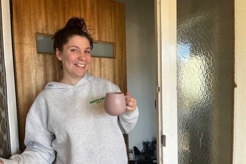 A smiling woman with dark hair in a bun wearing a grey hoodie holding a mug in the front doorway.