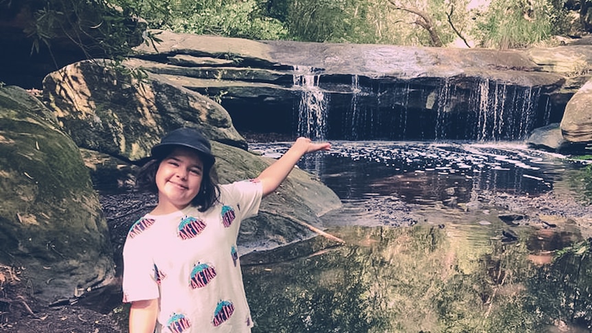 A girl stands by a natural stream with a bit of a waterfall behind her.