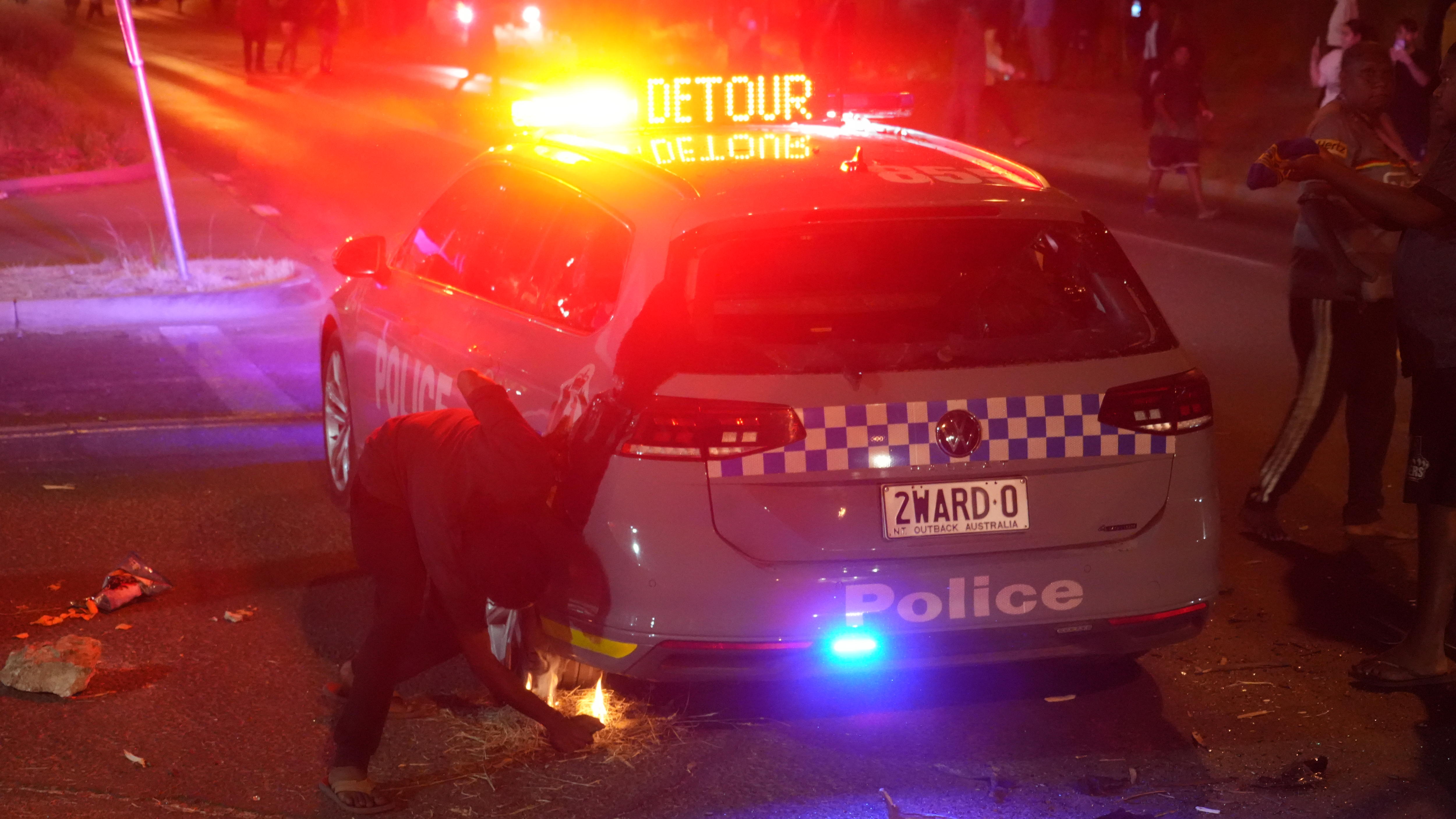 Un hombre intenta encender con un encendedor la hierba seca debajo de un vehículo policial por la noche.