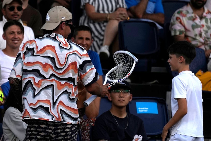 A fan in a crowd holds a damaged tennis racquet, with a younger spectator looking on