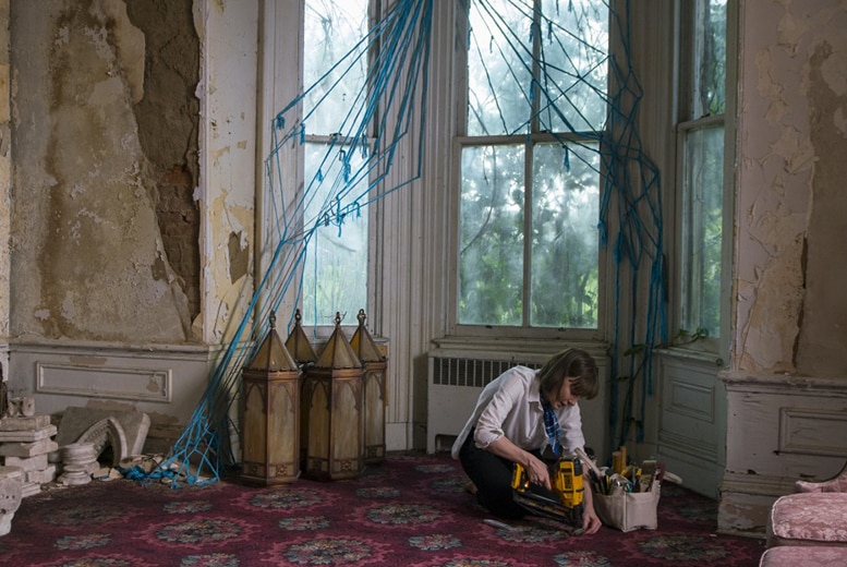 In the interior of a 1800s style dilapidated mansion mansion a woman is crouched down on colourful carpet with power tool.