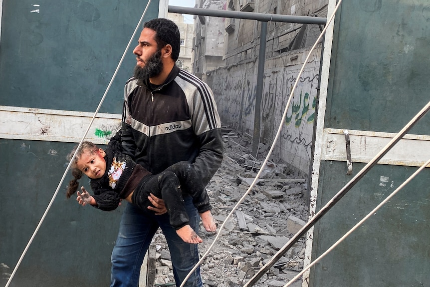 A man holding a young girl with a bloodied face walks out of a laneway lined with rubble of a destroyed building