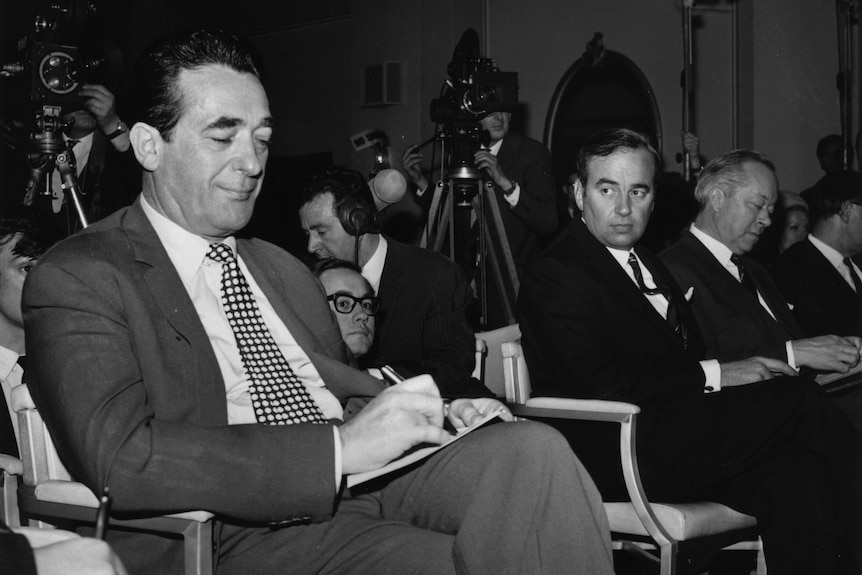 Robert Maxwell wears a suit while sitting and looking at his lap as Rupert Murdoch watches on from across the aisle.