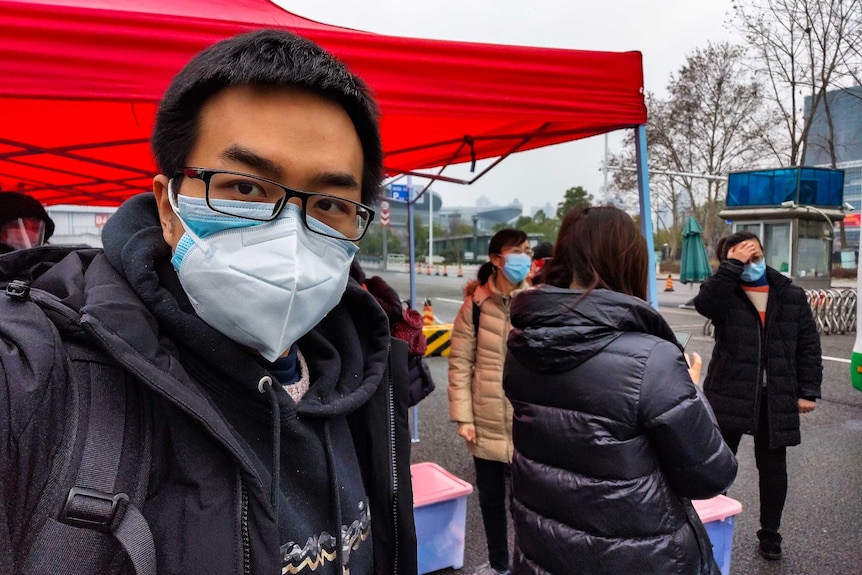A man with a face mask and glasses takes a selfie with a red marquee in the background.