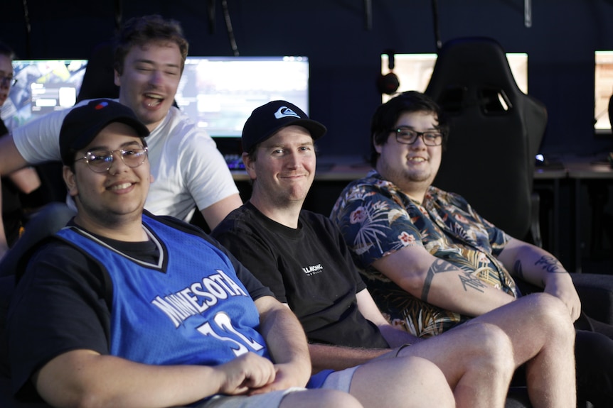 Three men sit together on a couch, while a young man in a white shirt photobombs them.