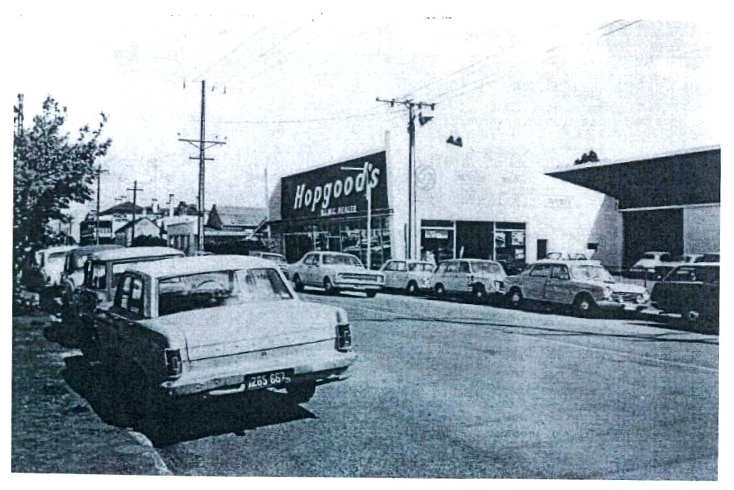 An old black and white photograph of a white commercial building with a big sign 'Hopgood's' above it. Cars line the street.