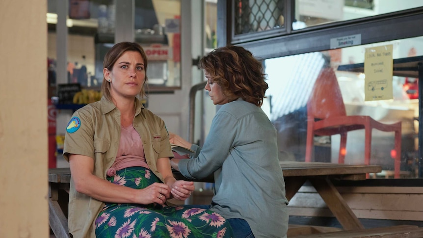 Brooke Satchwell sits on a bench facing the camera, looking concerned; Sigrid Thornton faces away from the camera