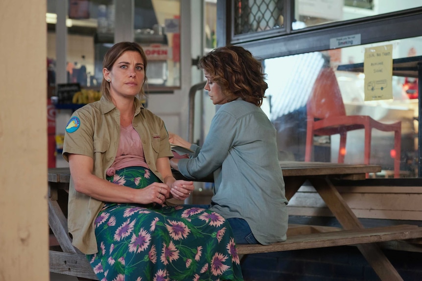 Brooke Satchwell sits on a bench facing the camera, looking concerned; Sigrid Thornton faces away from the camera
