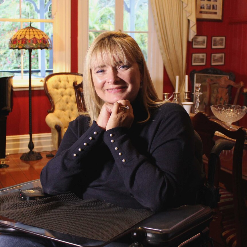 A woman is sitting in a wheelchair in her dining room with her hands held together under her chin.