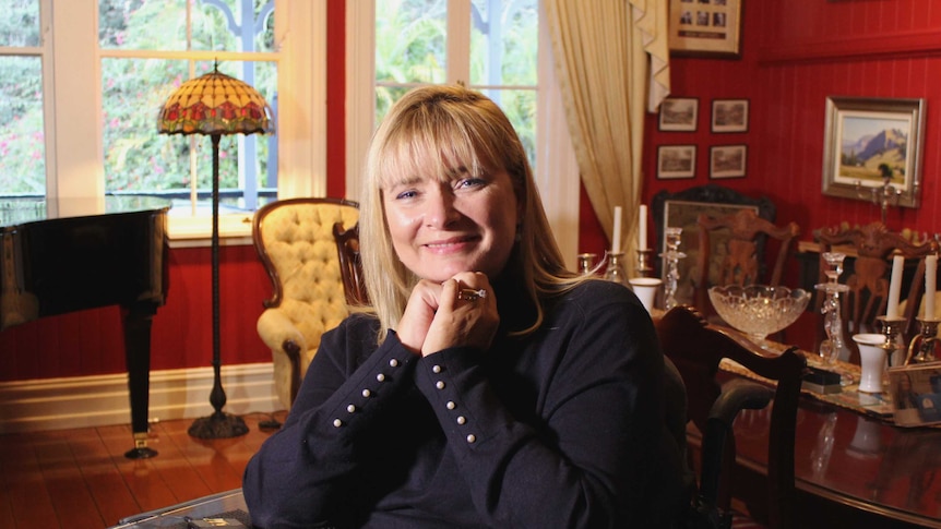 A woman is sitting in a wheelchair in her dining room with her hands held together under her chin.