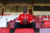 Young football player sits in the grandstand in his uniform and looks out towards the field.