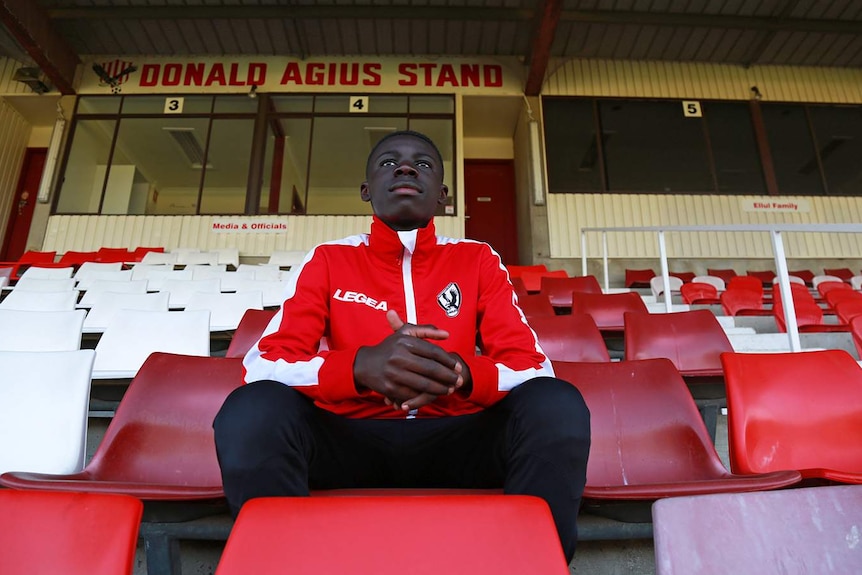 Young football player sits in the grandstand in his uniform and looks out towards the field.