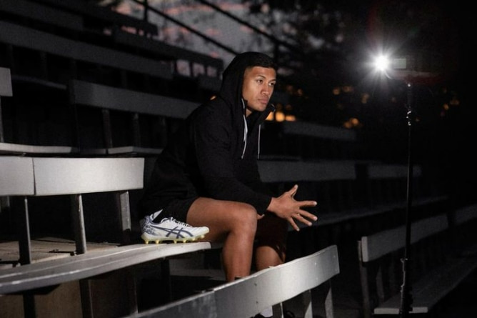 Man looks solemn as he sits in traning gear with football shoe beside him, at night, in the stands of a footy ground. 