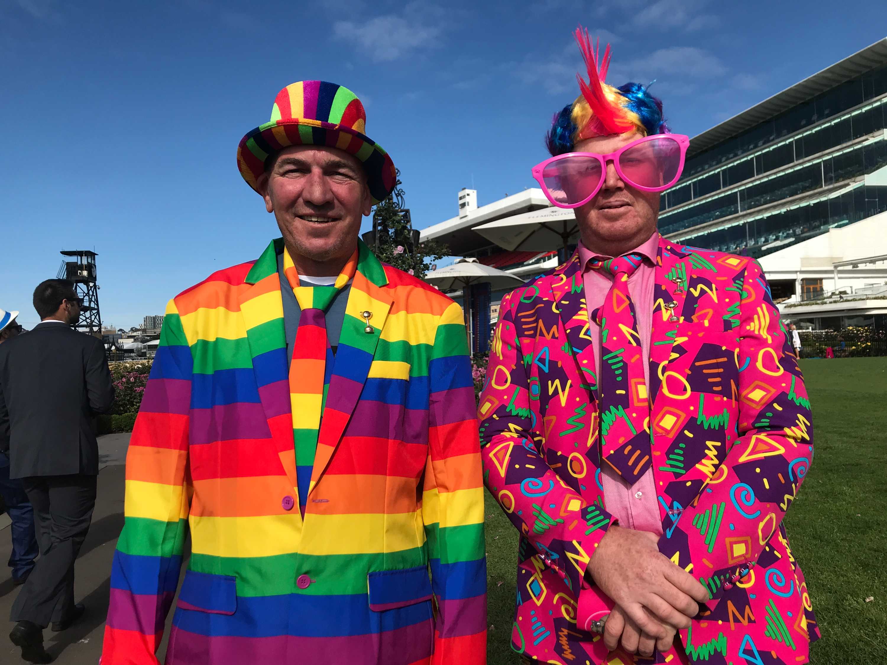 Two men dressed in multi-coloured suits stand in front on the grandstand at a race track.