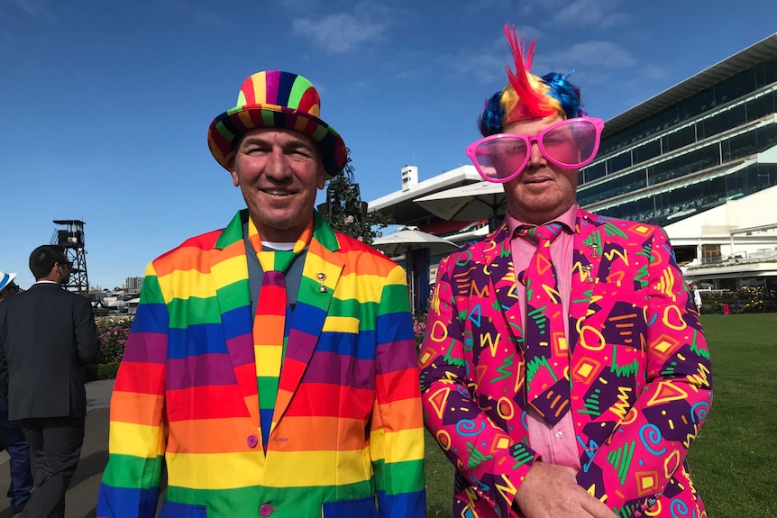 Two men dressed in multi-coloured suits stand in front on the grandstand at a race track.